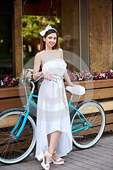 Beautiful girl in white dress posing with blue bike near a cafe.