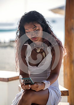 A beautiful girl in a white dress sits at a bar on the beach with a phone in her hands