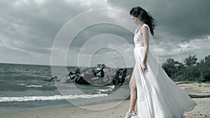 Beautiful girl in a white dress stands on seashore. Dress fluttering in wind slow motion.