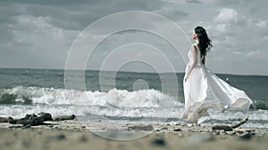 Beautiful girl in a white dress stands on seashore. Woman is holding a dress that flutters in wind slow motion.