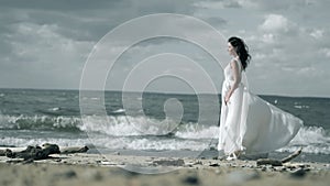 Beautiful girl in a white dress stands on seashore. Woman is holding a dress that flutters in wind slow motion.