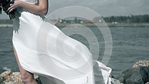 Beautiful girl in a white dress stands on seashore. Woman is holding a dress that flutters in wind slow motion.