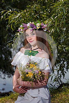 Girl in white dress and wreath holding field flowers bouquet on green willow background. 7th July, traditional slavic holiday with