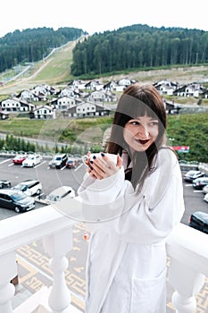 Beautiful girl in white hotel bathrobe drinking hot tea or coffee on a balcony. Nature and nice view outside. Quiet weekend