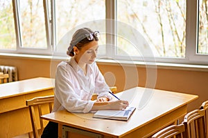 Beautiful girl in a white shirt sitting at the table by the window in the classroom reading a book