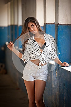 Beautiful girl with white shirt and white shorts posing in old hall with columns blue painted. Attractive long hair brunette