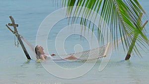 A beautiful girl in a white swimsuit relaxes while lying in a hammock and drinks a coconut. Beach holiday luxury concept