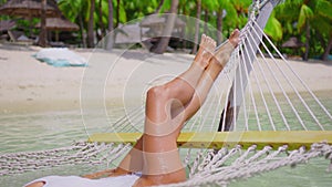 A beautiful girl in a white swimsuit relaxes while lying in a hammock and drinks a coconut. Beach holiday luxury concept