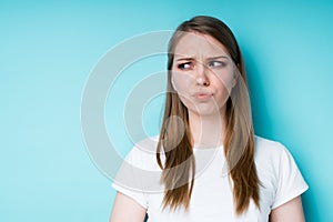 A beautiful girl in a white T-shirt pouts her lips offendedly and looks to the side while standing on a blue background