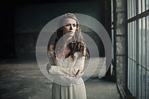 Beautiful girl in in white vintage dress with curly hair posing near the loft window. Woman in retro dress. Worried sensual emotio