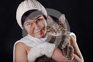 Beautiful girl in a white woolen hat and scarf with a tabby cat on black background