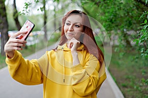 A beautiful girl in a yellow hoodie takes a selfie, holds her phone in her hands and adjusts her hair