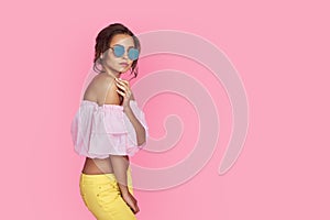 Beautiful girl in yellow jeans and pink shirt, with hands up wearing sunglasses posing, dancing, smiling on pink