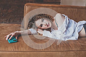 Beautiful glamorous woman in white shirt with curly hair lies on the sofa with green mobile phone in her hand