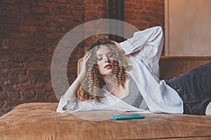 Beautiful glamorous woman in white shirt with curly hair lies on the sofa with green mobile phone, touching her hair