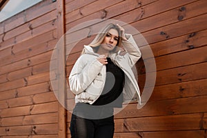 Beautiful glamour model of a young woman with blond long hair in stylish black clothes in a fashionable white jacket poses near a