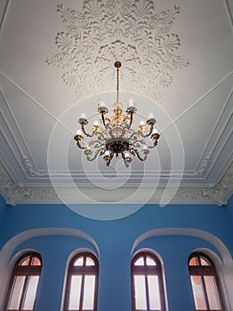 Beautiful golden chandelier suspended down a white ornate ceiling inside a palace room with blue walls and arched windows.