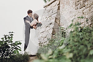 Beautiful gorgeous bride and groom walking in sunny park and kissing. happy wedding couple hugging in green garden at old castle.