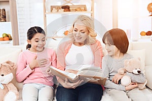 Beautiful grandmother looks at photo album with joyful granddaughter and grandson holding teddy bear.