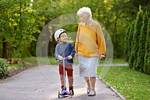 Beautiful granny and her little grandchild walking together in autumn park. Boy riding by scooter