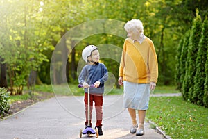 Beautiful granny and her little grandchild walking together in autumn park. Boy riding by scooter