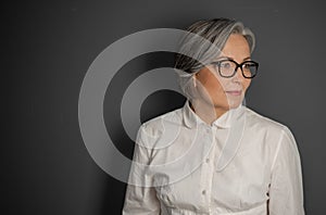Beautiful gray-haired business woman looking at side. Mature woman in glasses and white shirt on grey background
