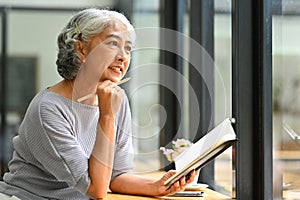 Beautiful gray haired mature woman holding book and looking away, daydreaming or thinking on good retirement plans