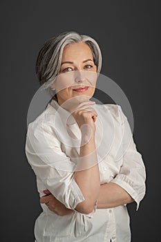 Beautiful gray-haired woman gently smiles while touching her chin with hand and looking at camera. Intelligent beautiful