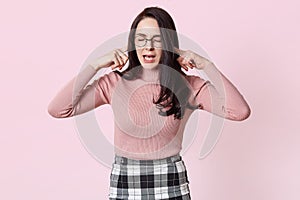 Beautiful grimacing young woman in pink sweater, her ears with fingers and she keeps eyes closed. Charming female with long dark