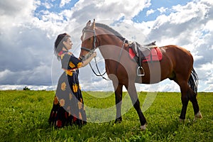 Beautiful gypsy girl with a horse in field with green glass in summer day and blue sky and white clouds background. Model in