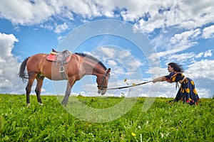 Beautiful gypsy girl leads a stubborn, unruly horse in field with green glass in summer day and blue sky and white clouds