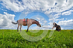 Beautiful gypsy girl leads a stubborn, unruly horse in field with green glass in summer day and blue sky and white clouds