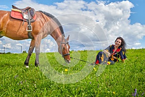 Beautiful gypsy girl sitting near horse in field with green glass in summer day and blue sky and white clouds background. Model in