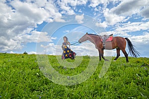 Beautiful gypsy girl walking with a horse in field with green glass in summer day and blue sky and white clouds background. Model