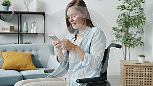 Beautiful handicapped woman using smart phone texting and smiling sitting in wheelchair in apartment