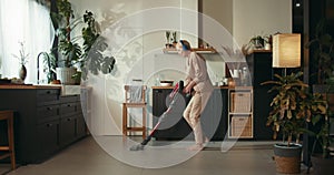 Beautiful happy 60s senior woman having fun dancing to music in headphones, cleaning floor with cordless vacuum cleaner.