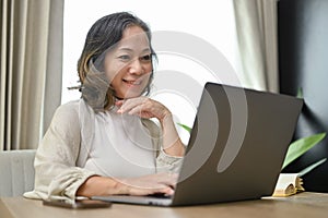 Beautiful and happy Asian-aged woman using laptop in her living room, browsing internet