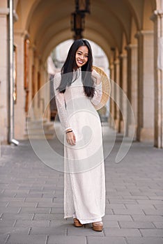 Beautiful happy asian girl dressed in Ao Dai dress with vietnamese conical hat against arches background.