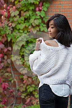 Beautiful happy Asian girl looks away in white knitted cardigan near brick wall in a street