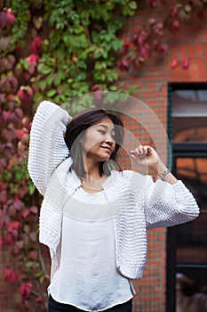 Beautiful happy Asian girl looks away in white knitted cardigan near brick wall in a street