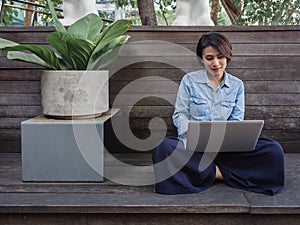 Beautiful happy asian woman wearing blue jeans shirt using laptop computer on her lap on wooden background