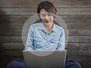 Beautiful happy asian woman wearing blue jeans shirt using laptop computer on her lap on wooden background