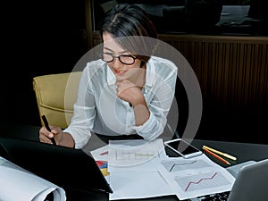 Beautiful happy Asian woman working overtime with laptop computer on desk in office