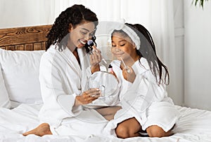 Beautiful happy black mother and little daughter in bathrobes applying makeup together