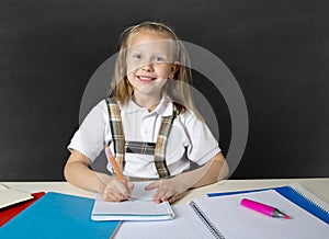 Beautiful happy blond junior schoolgirl smiling while doing school homework writing on notepad with pen