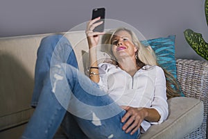 Beautiful happy blond woman early 40s relaxed at home living room using internet social media on mobile phone smiling lying comfor