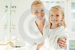 beautiful happy blonde mother and daughter in bathrobes smiling at camera