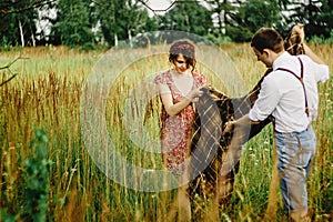 beautiful happy bride and stylish retro groom holding tweed blanket or plaid, picnic time in sunny meadow