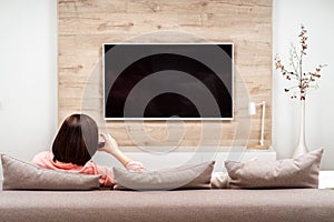 Beautiful happy brown-haired young female relaxing with glass of red wine on the sofa and watching tv.