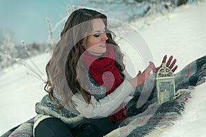 Beautiful happy brunette girl in a red knitted scarf sitting on a blanket on the snow in winter on background of trees